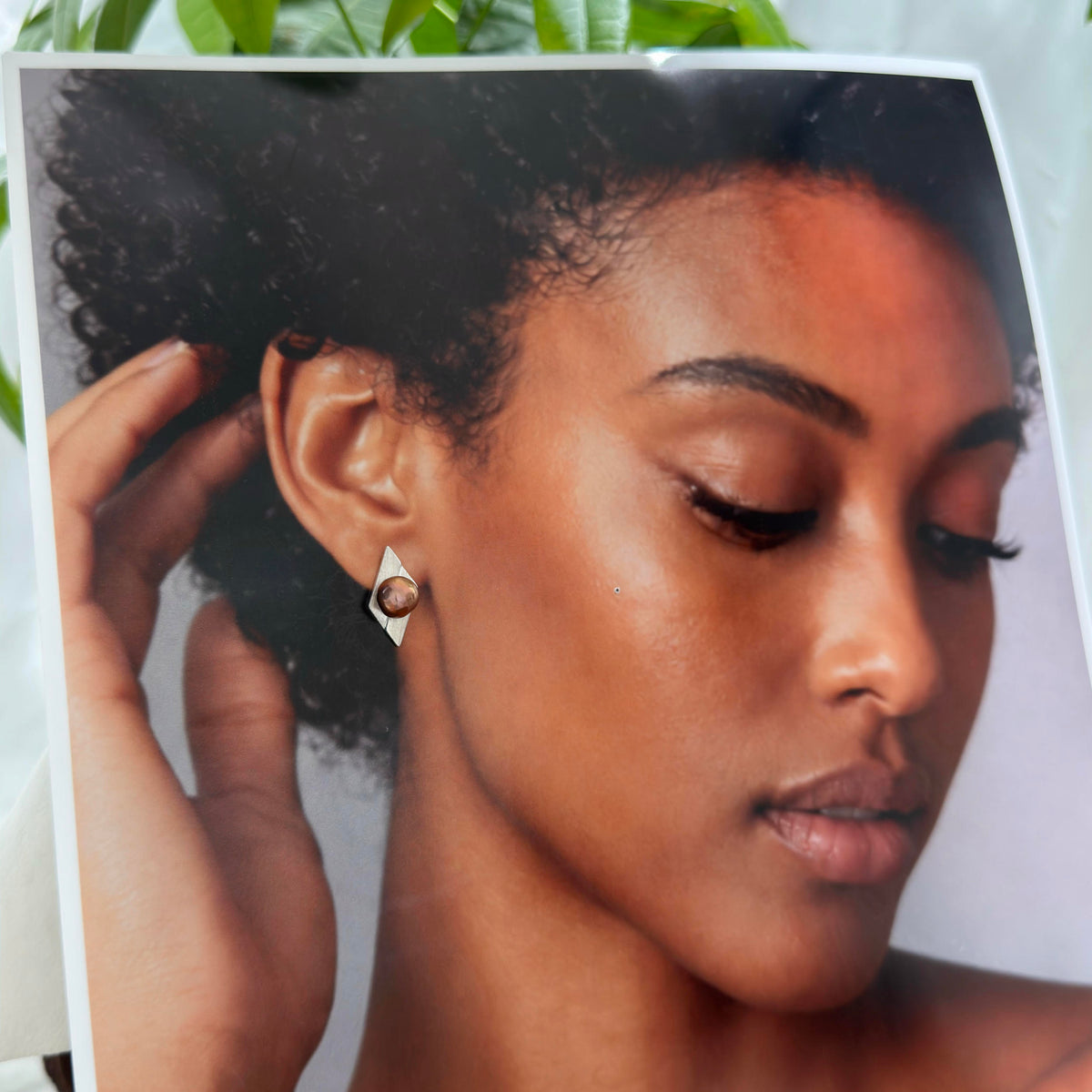 A close-up portrait of a woman with dark curly hair wearing a small geometric stud earring, shown within a printed or framed photo that is partially visible against a light background with green leaves at the top.
