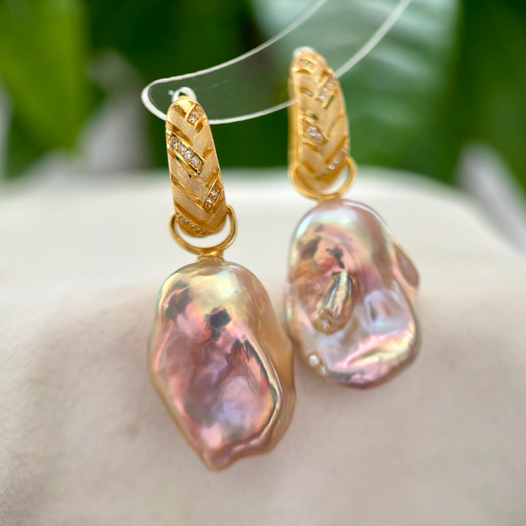 A pair of gold hoop earrings with baroque pearl drops, displayed on a clear stand against a soft-focus background of green leaves and a light beige surface.