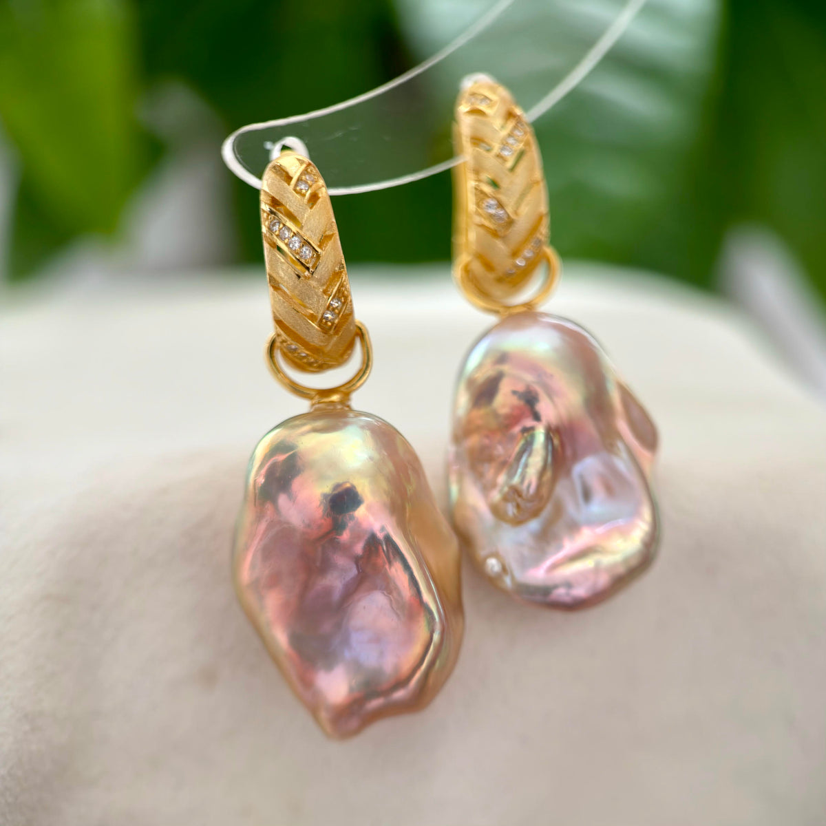 A pair of gold hoop earrings with baroque pearl drops, displayed on a clear stand against a soft-focus background of green leaves and a light beige surface.