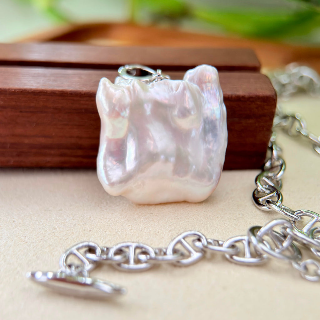 A close-up of a silver chain necklace with a large, irregularly shaped white pearl pendant resting on a wooden block, with the silver clasp chain visible in the foreground and background.