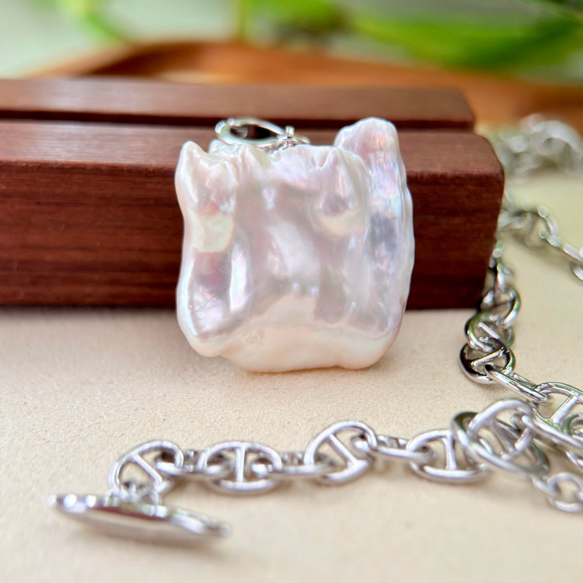 A close-up of a silver chain necklace with a large, irregularly shaped white pearl pendant resting on a wooden block, with the silver clasp chain visible in the foreground and background.
