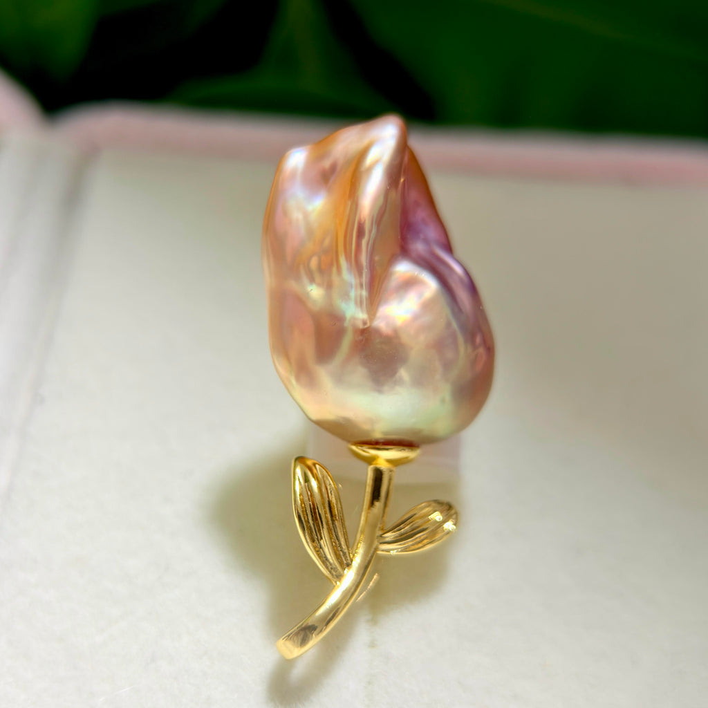 A baroque pearl rose brooch with a gold metal stem and leaves, displayed on a white textured surface.