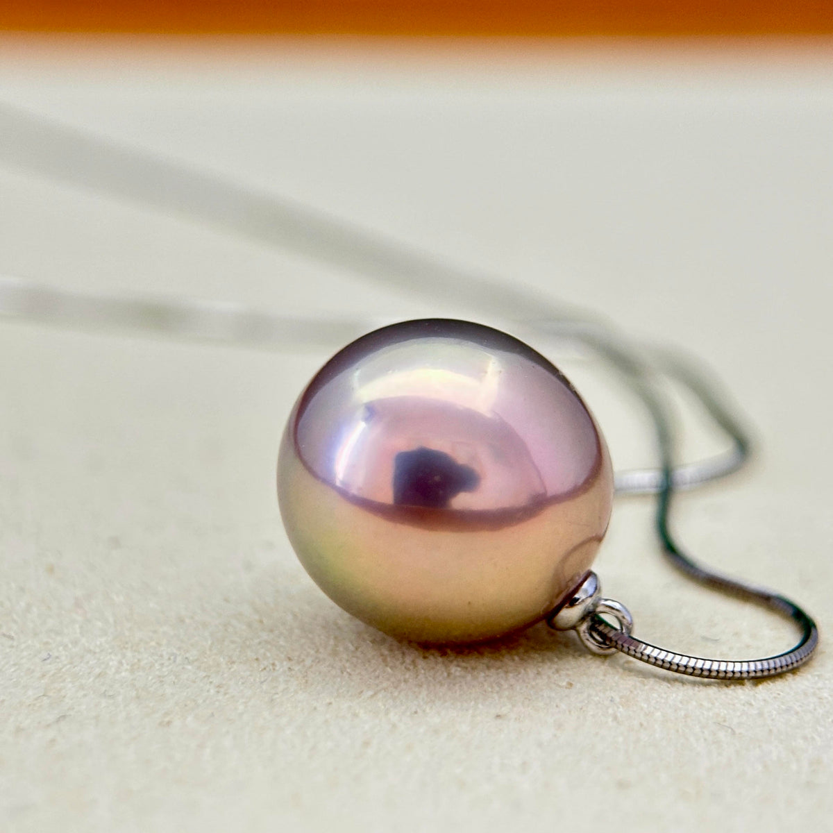 A close-up of a round iridescent pearl pendant necklace on a silver-toned chain, resting on a light beige textured surface.