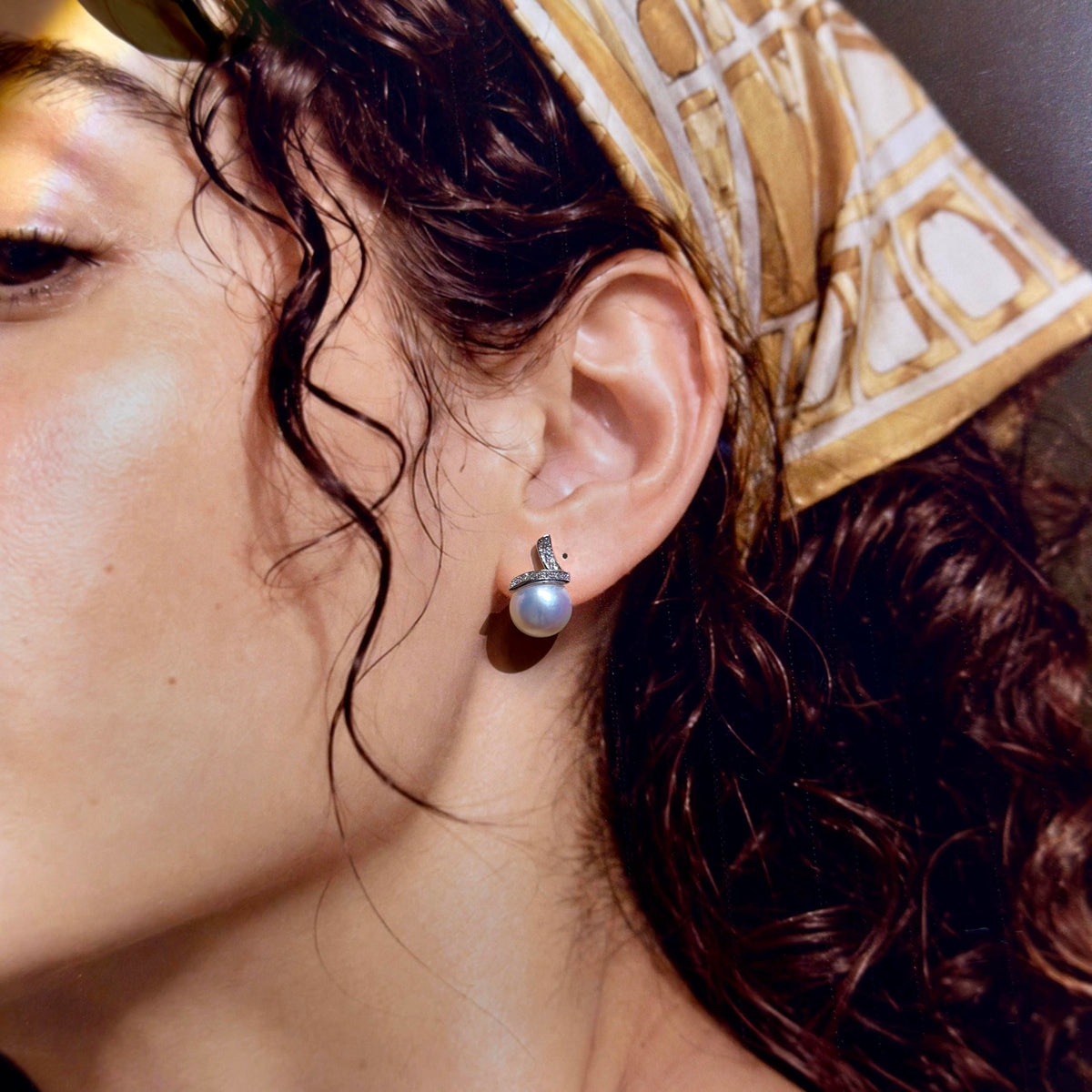 A close-up of a woman's ear wearing a silver stud earring with a single round white pearl, with her dark curly hair and a gold and white patterned headscarf visible.