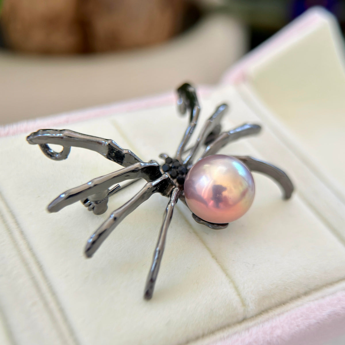 A spider-shaped brooch with a glossy iridescent pink pearl and dark metallic legs, displayed on a cream-colored jewelry box.