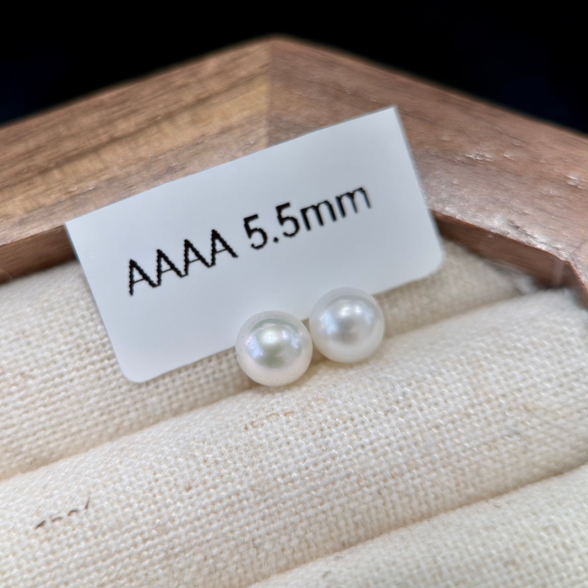 Two round white pearl stud earrings resting on a beige fabric jewelry display, with a small white card above them reading 'AAAA 5.5mm'.