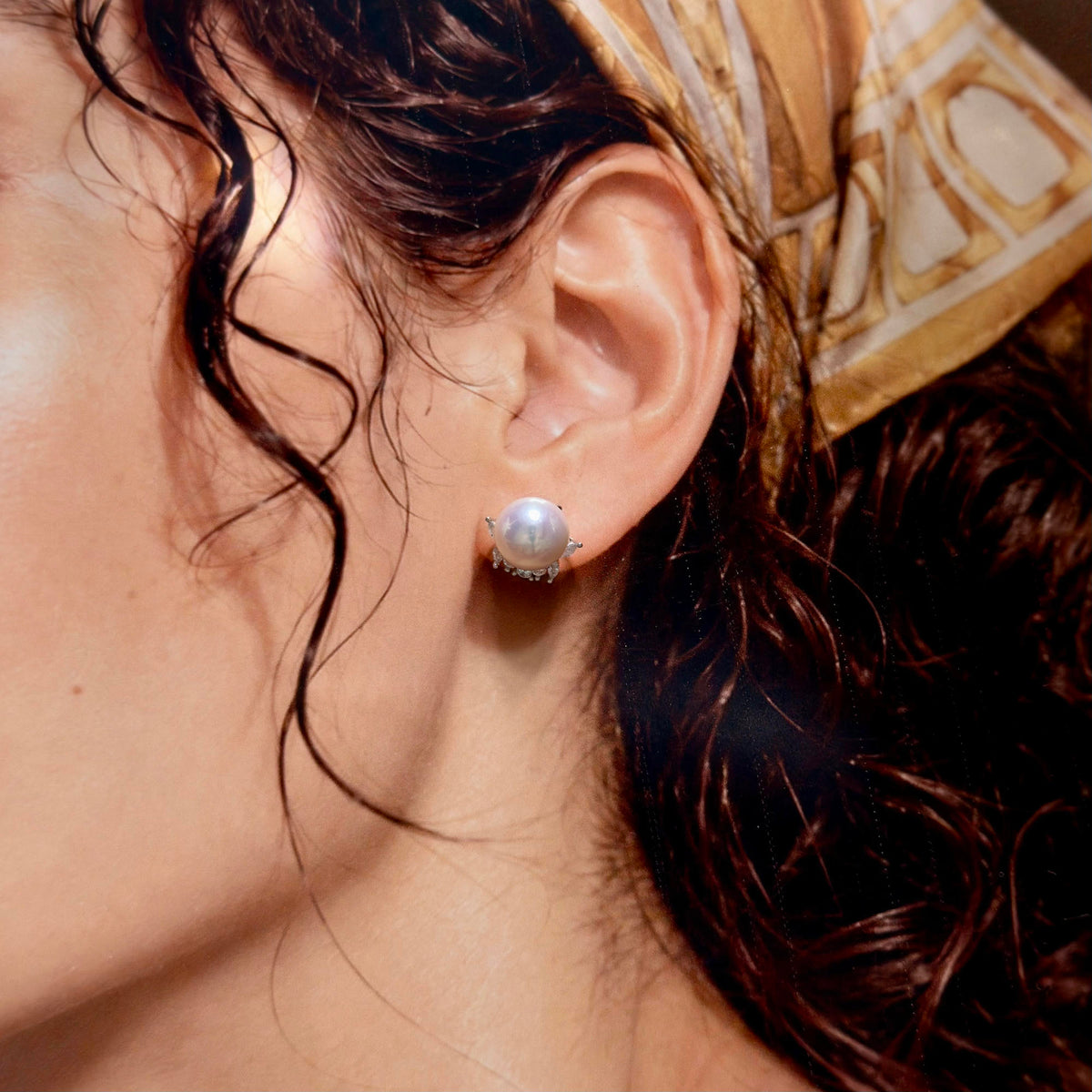 A close-up of a woman's ear wearing a single pearl stud earring with a silver-toned setting and small clear accent stones.