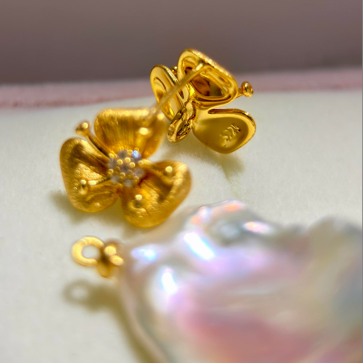 A close-up of a gold flower-shaped pendant or charm with a small iridescent pearl-like center, shown on a soft cream surface with a blurred iridescent pearl in the foreground.