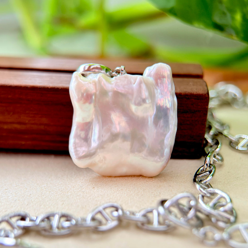 A close-up of a chunky, irregularly shaped iridescent white pearl pendant on a silver link chain, resting on a wooden block with a soft-focus green background.