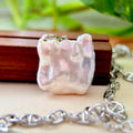 A close-up of a chunky, irregularly shaped iridescent white pearl pendant on a silver link chain, resting on a wooden block with a soft-focus green background.