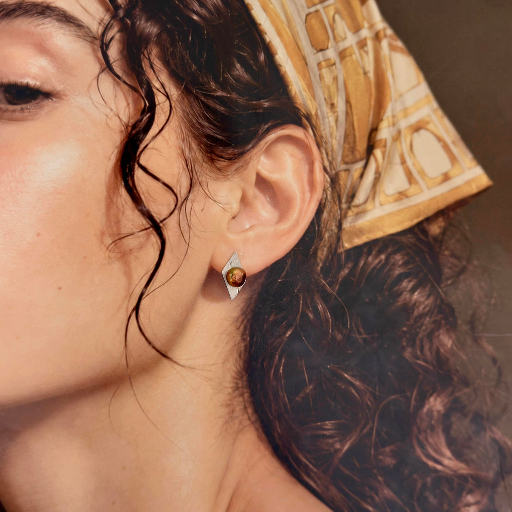 A close-up of a woman's ear wearing a small silver geometric stud earring with a round amber-brown stone, against warm skin tones and a patterned headscarf.