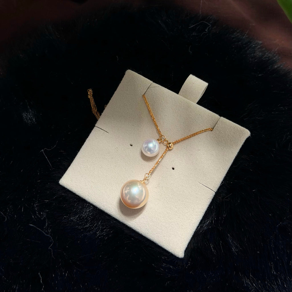 A gold necklace with two pearl-like drops, one larger and one smaller, displayed on a cream jewelry card over a black furry surface.