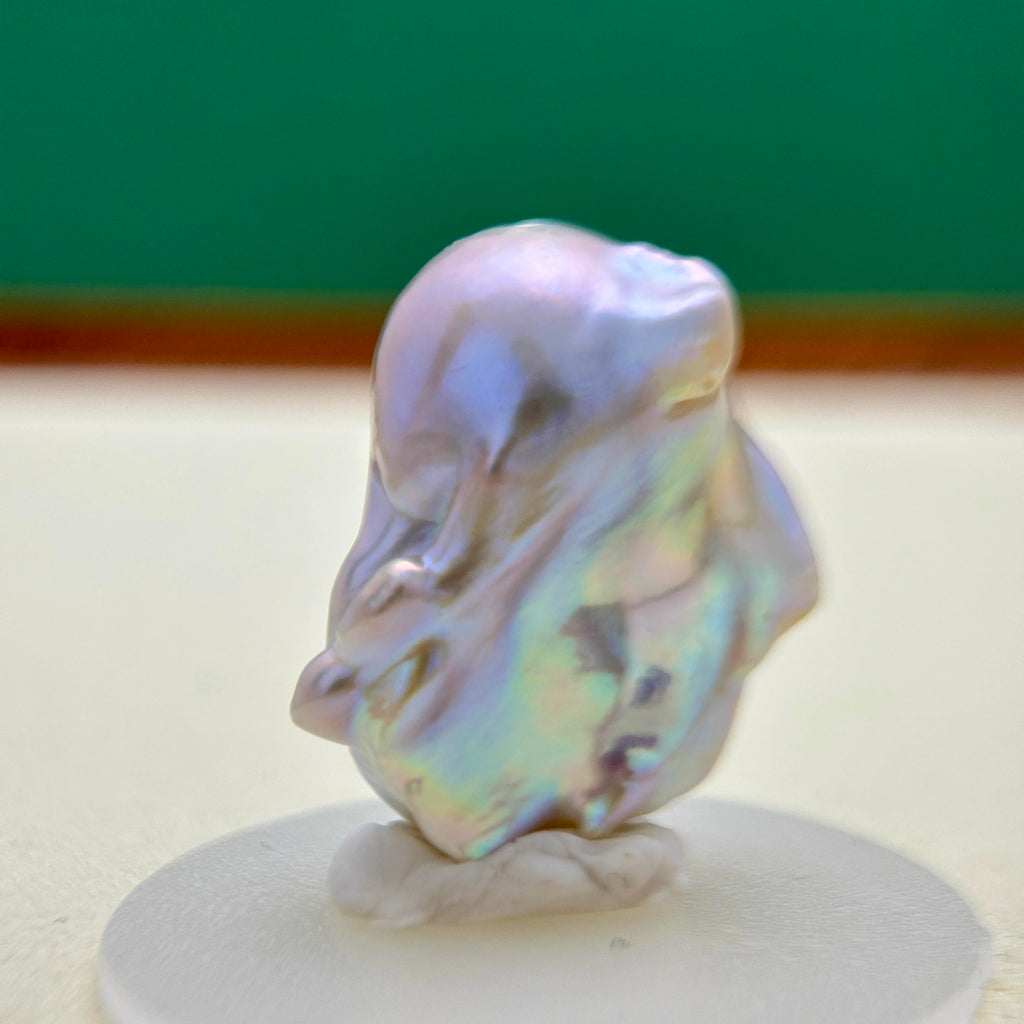 A baroque pearl with an iridescent, pearly luster showing shades of lavender, blue, and green, displayed on a small white stand against a blurred background.