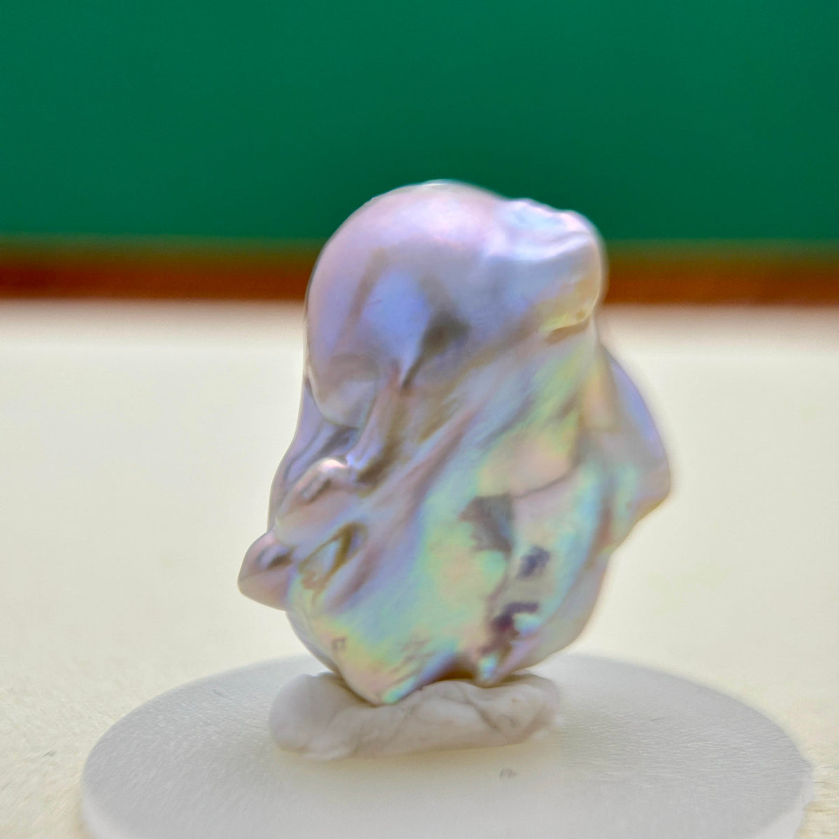 A baroque pearl with an iridescent, pearly luster showing shades of lavender, blue, and green, displayed on a small white stand against a blurred background.