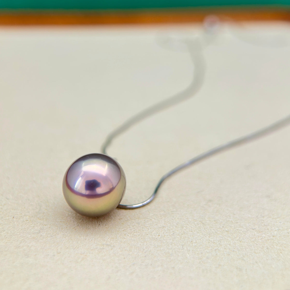 A single round freshwater pearl pendant with a pearlescent lavender-purple and green iridescence, suspended on a thin silver wire necklace against a neutral beige background.