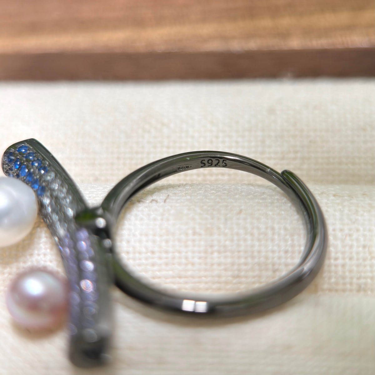 A close-up of a dark metallic open ring with a curved bar set with blue and clear stones, resting on a textured cream fabric surface with a wooden background.