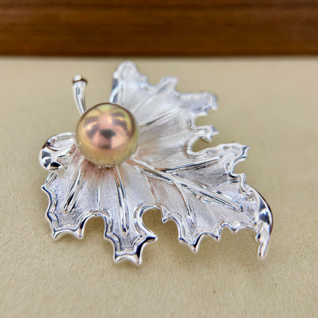 A silver-toned leaf-shaped brooch with a central iridescent pearl, displayed on a beige surface.