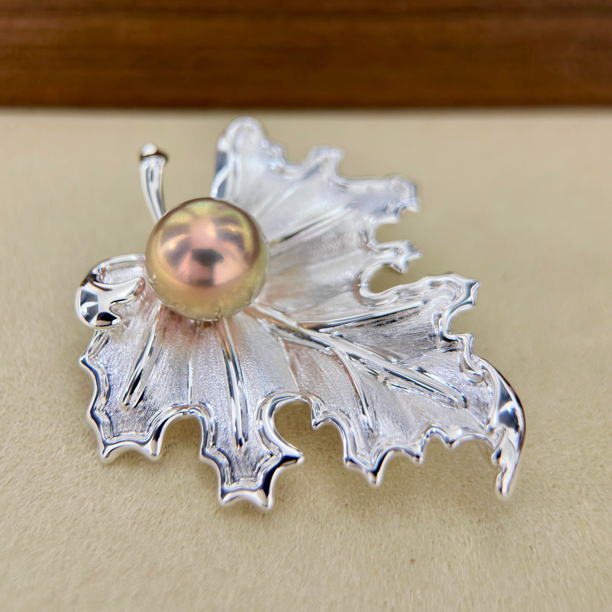 A silver-toned leaf-shaped brooch with a central iridescent pearl, displayed on a beige surface.