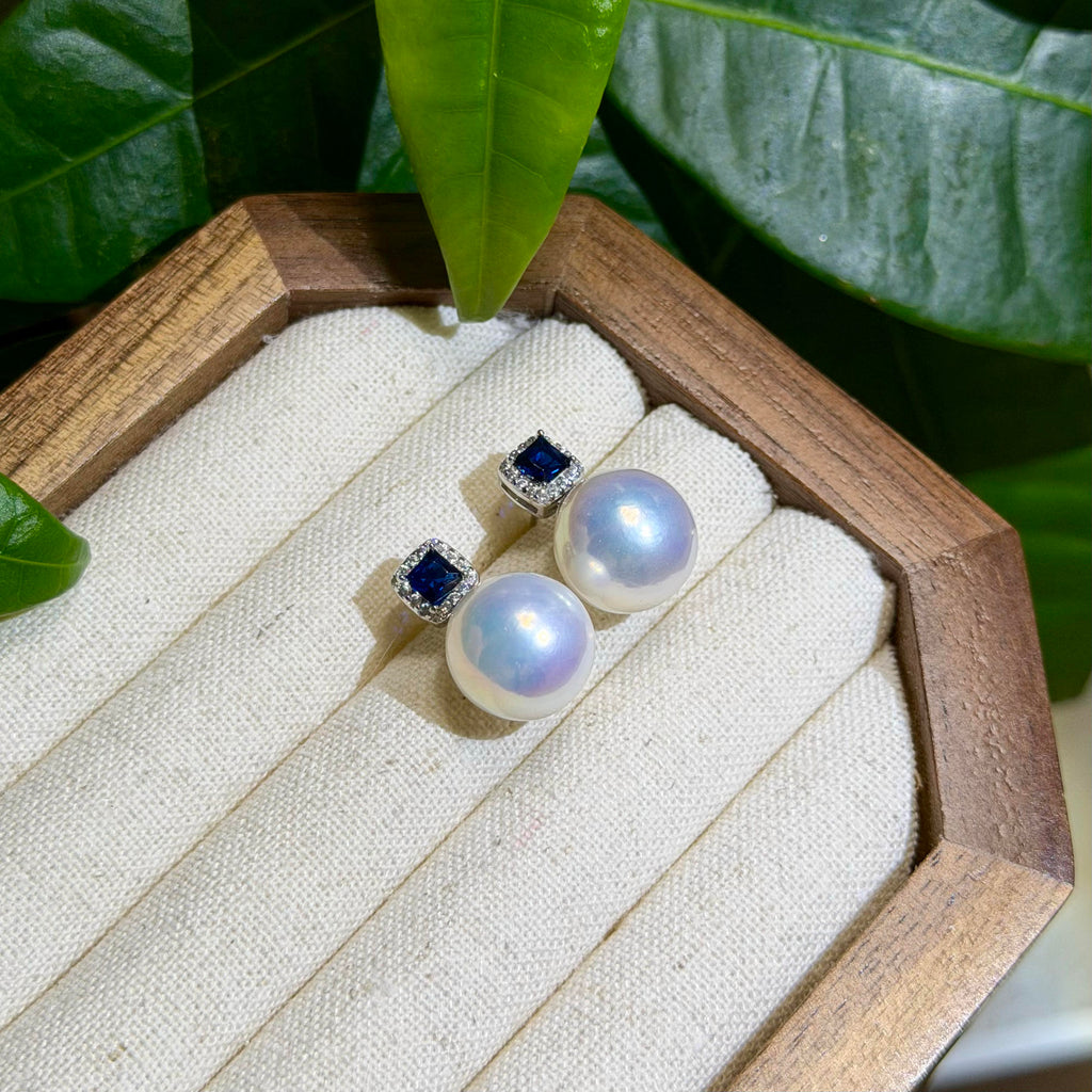 A pair of stud earrings displayed on a cream fabric jewelry tray inside a wooden octagonal box, with large green leaves in the background. The earrings feature round white pearls and square dark blue stones in silver settings.
