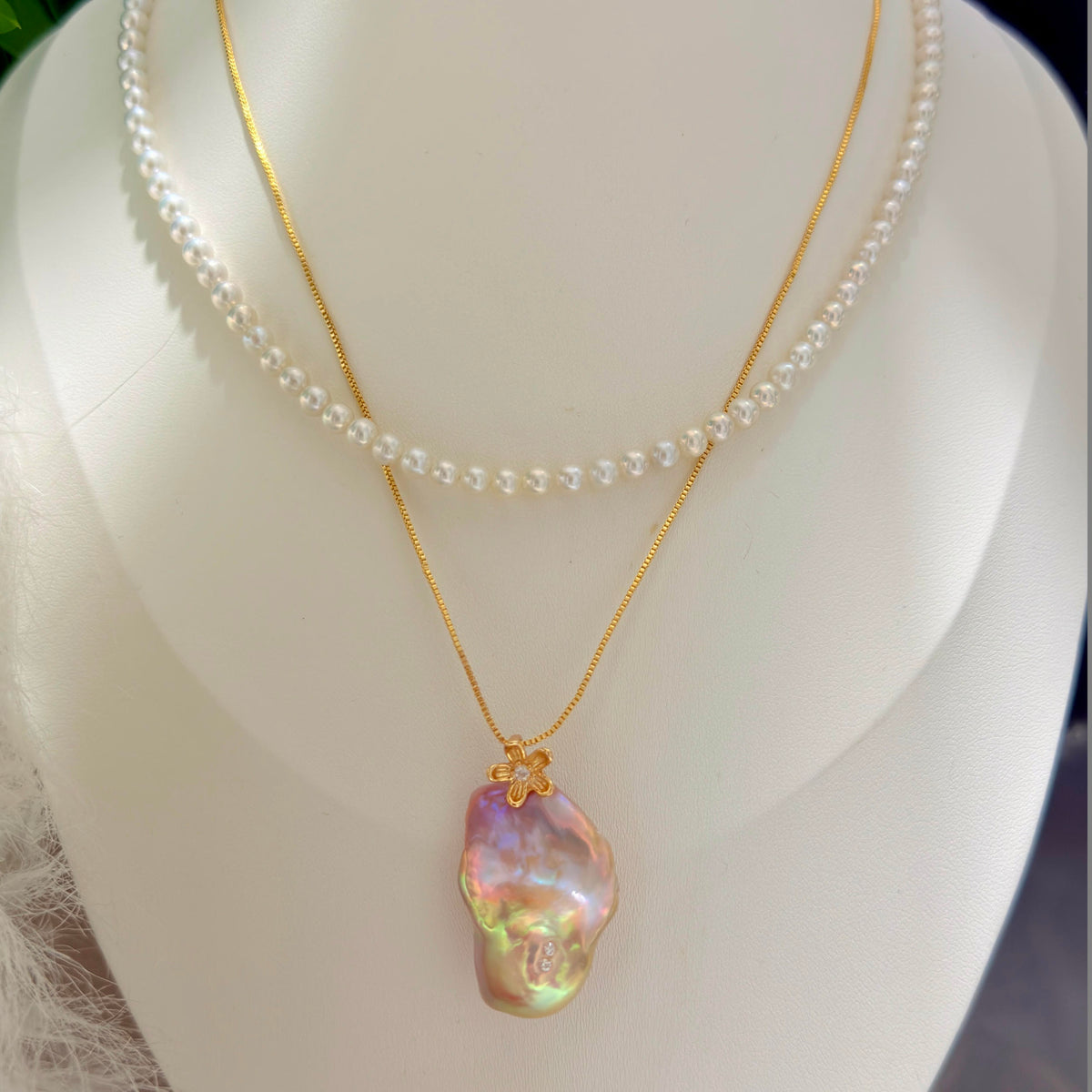 A double-strand necklace displayed on a white mannequin bust, featuring a delicate white pearl strand and a gold chain with an irregular, iridescent baroque pearl pendant.