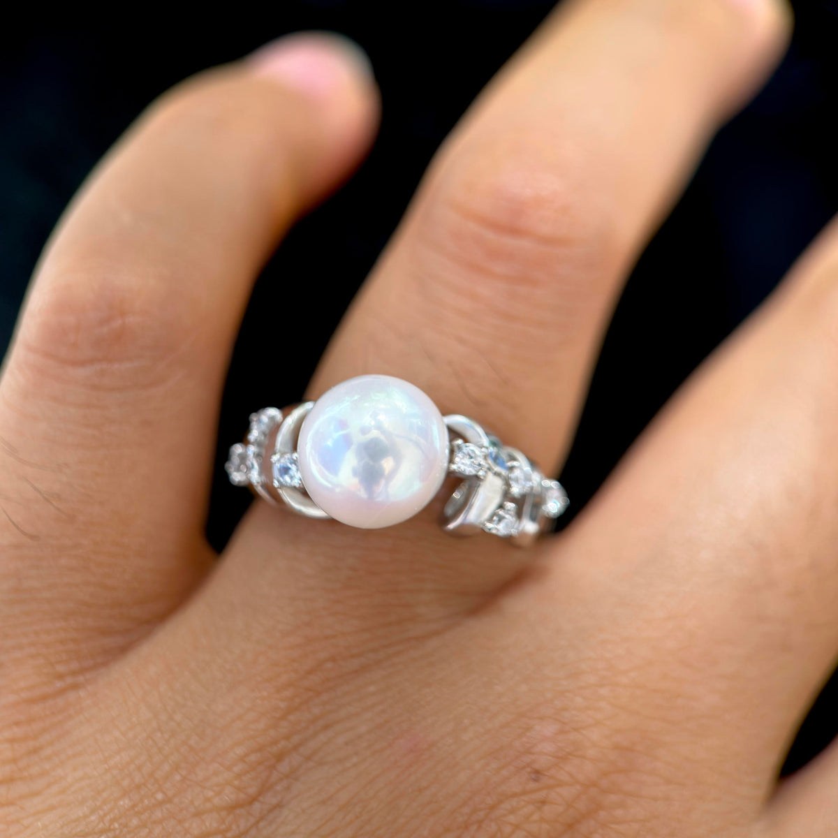 A silver ring featuring a large, round, lustrous white pearl set with small clear stones on a finger, shown against a dark background.