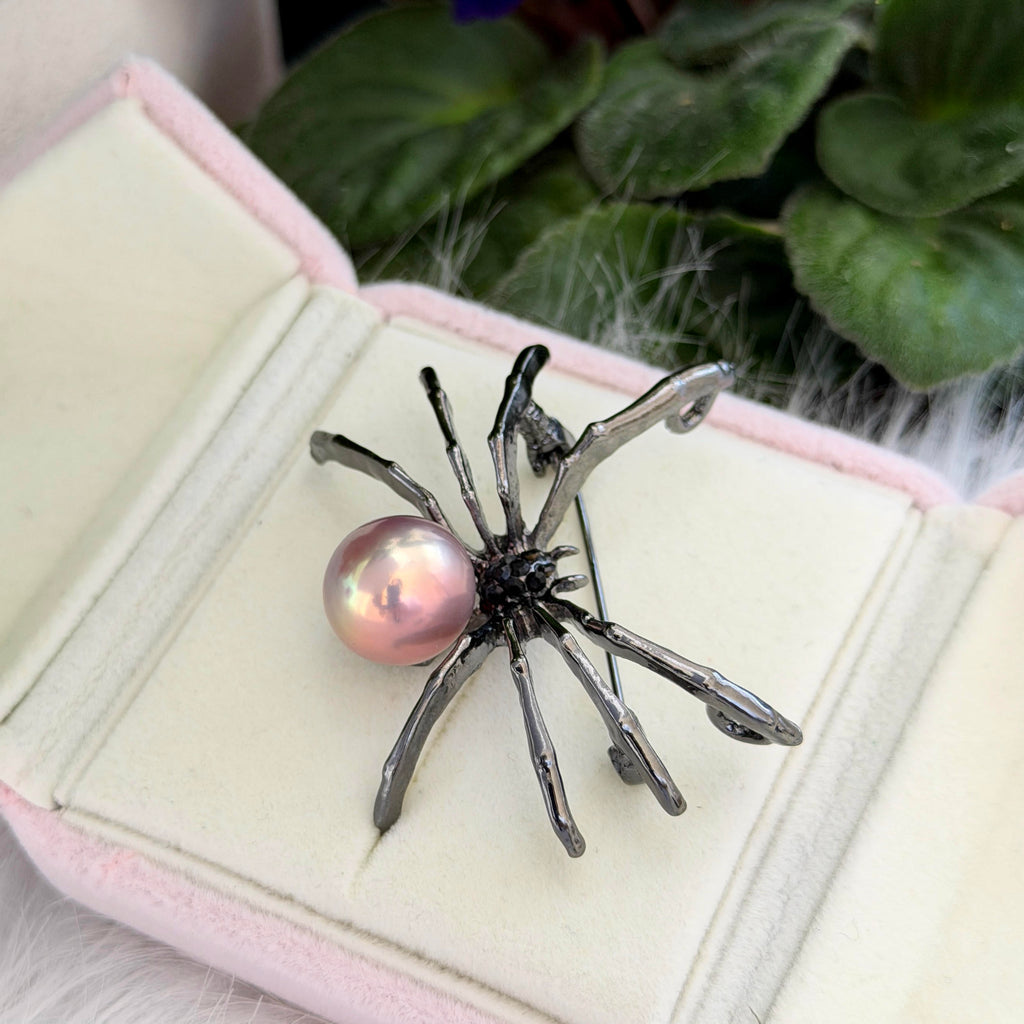 A spider-shaped brooch with a glossy pink pearl and dark metallic legs, displayed inside an open pale pink jewelry box with green leaves in the background.