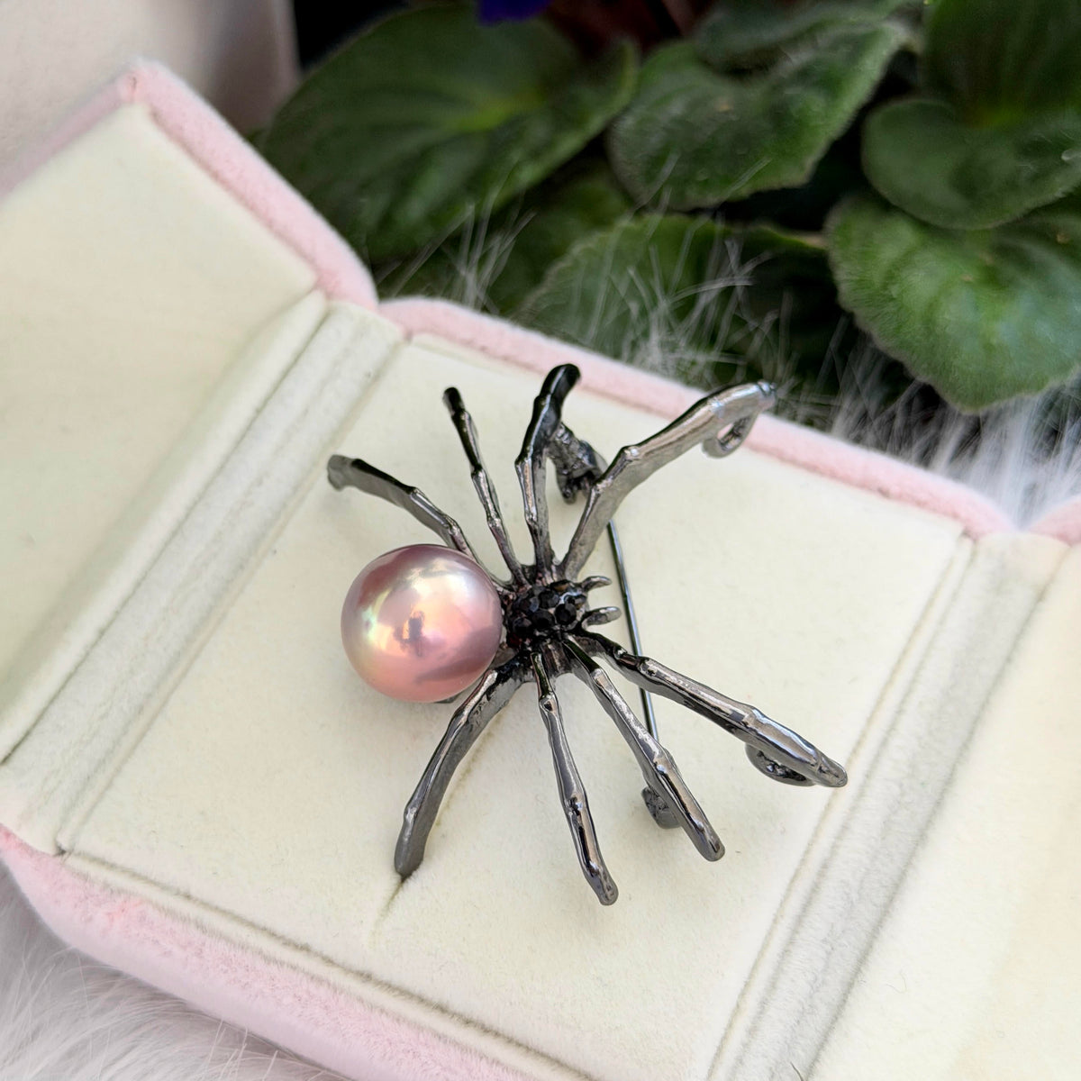 A spider-shaped brooch with a glossy pink pearl and dark metallic legs, displayed inside an open pale pink jewelry box with green leaves in the background.