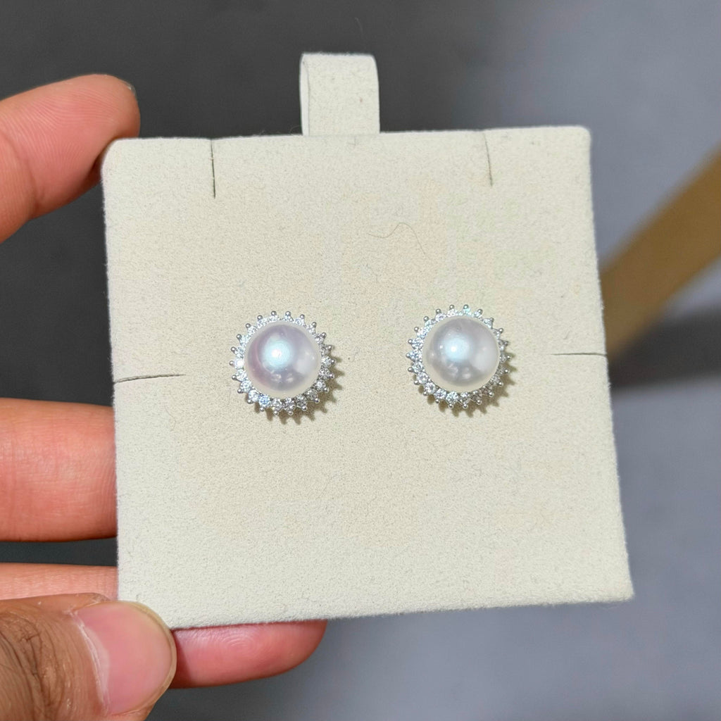 A pair of pearl stud earrings with silver halo settings, displayed on a beige jewelry card held by a hand.