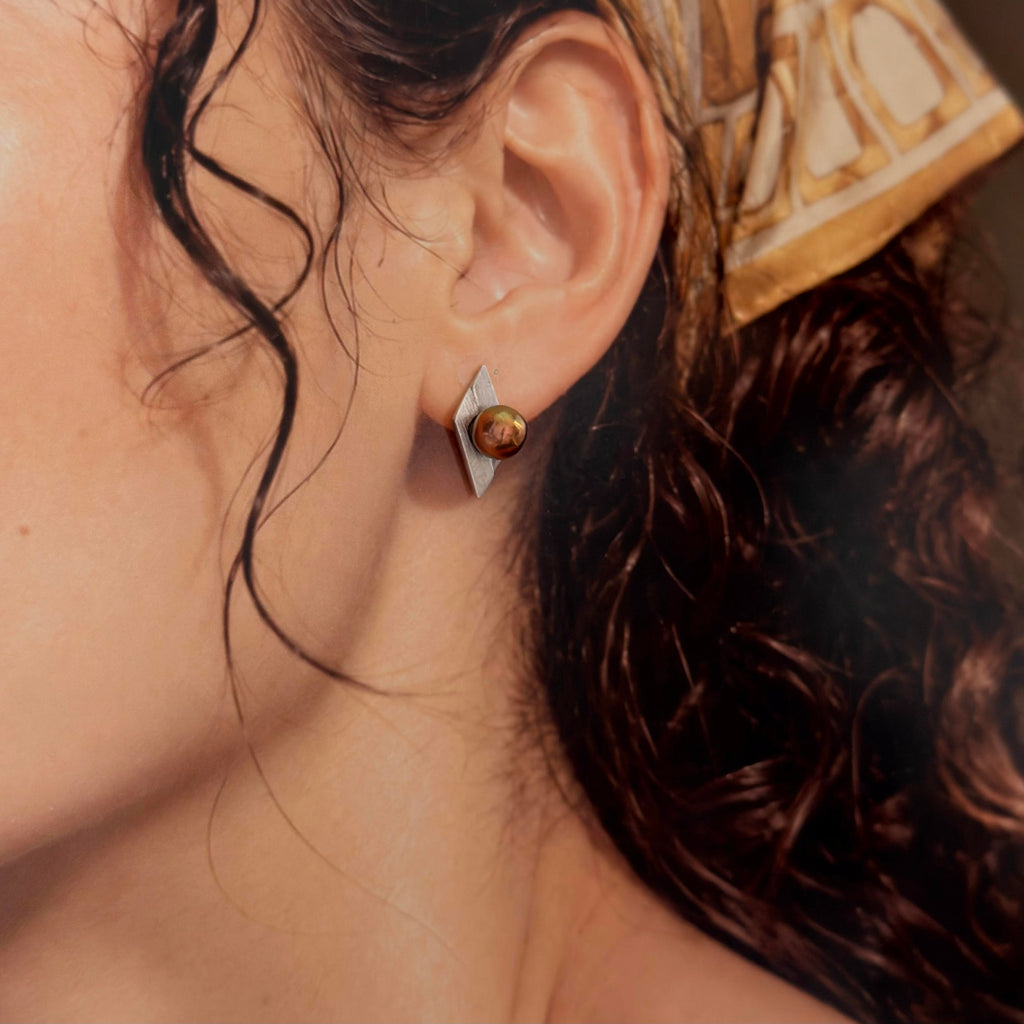 A close-up of a person's ear wearing a geometric silver stud earring with a round amber-brown center, against warm skin tones and dark hair.