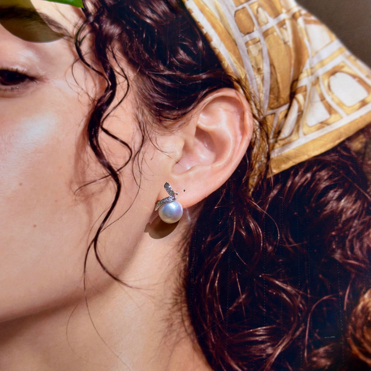 A close-up of a person's ear wearing a silver stud earring with a single white pearl drop, against a background of dark brown hair and a gold patterned headscarf.