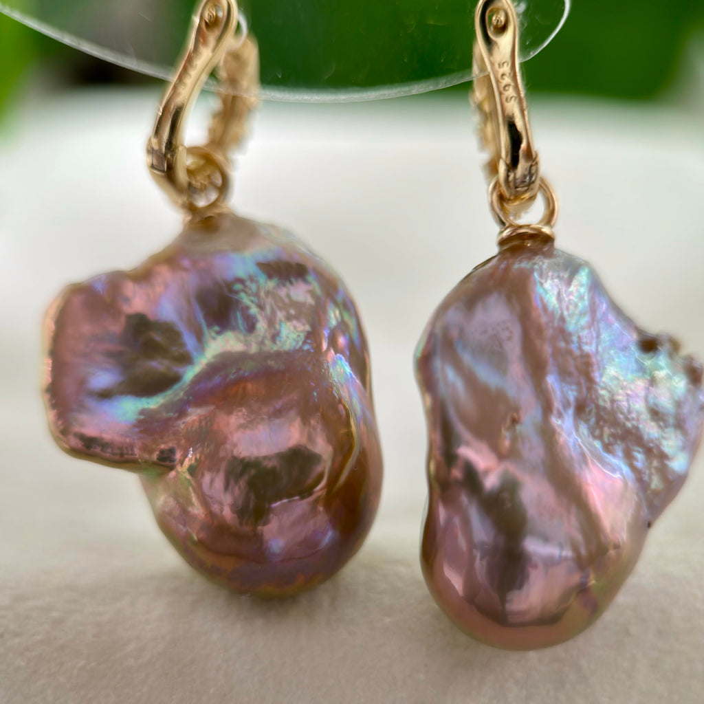 A pair of gold hoop earrings with large, organic baroque pearl drops that shimmer with iridescent pink, purple, and green hues, resting on a soft white surface.