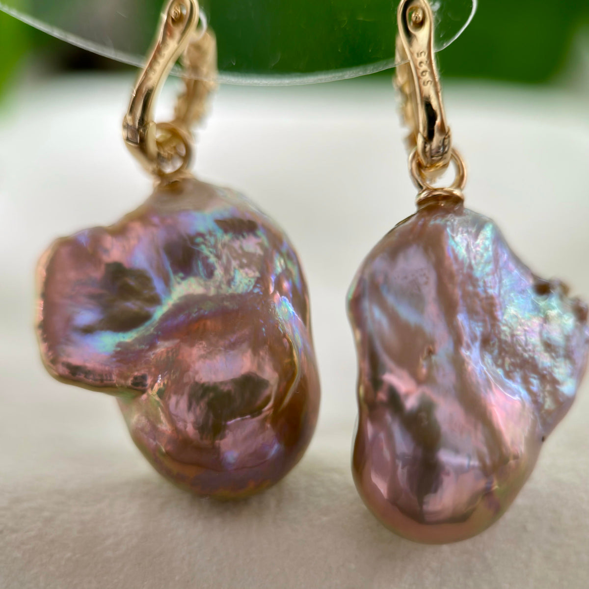 A pair of gold hoop earrings with large, organic baroque pearl drops that shimmer with iridescent pink, purple, and green hues, resting on a soft white surface.