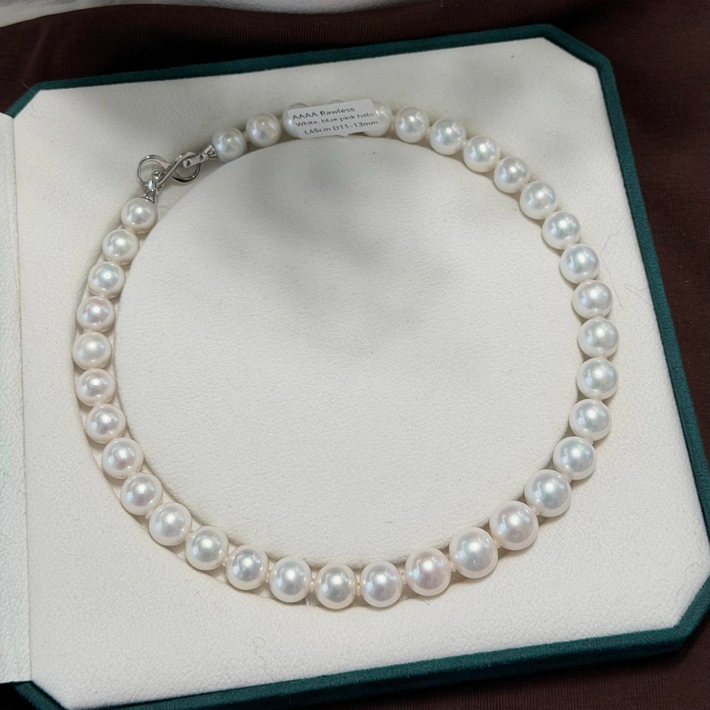 A pearl necklace arranged in a white jewelry box insert, with a small silver clasp visible at the top. The box is dark green-edged and placed on a dark brown surface.