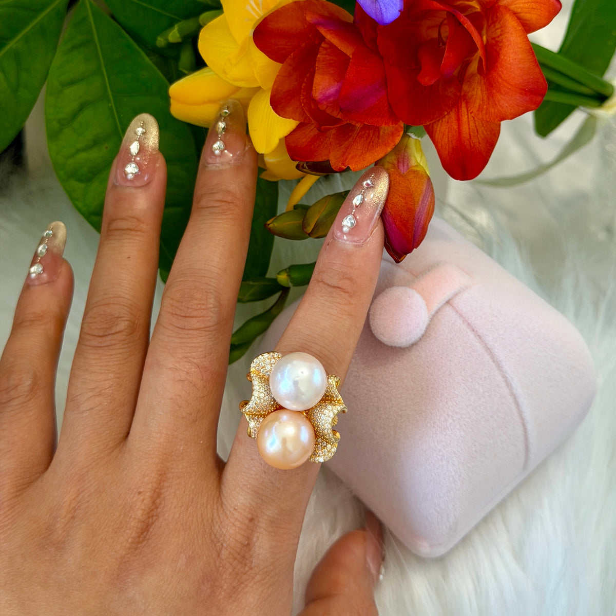 A hand with decorated nails wears a gold ring featuring two large white pearls and small clear rhinestones. A pale pink ring box and colorful flowers are in the background.