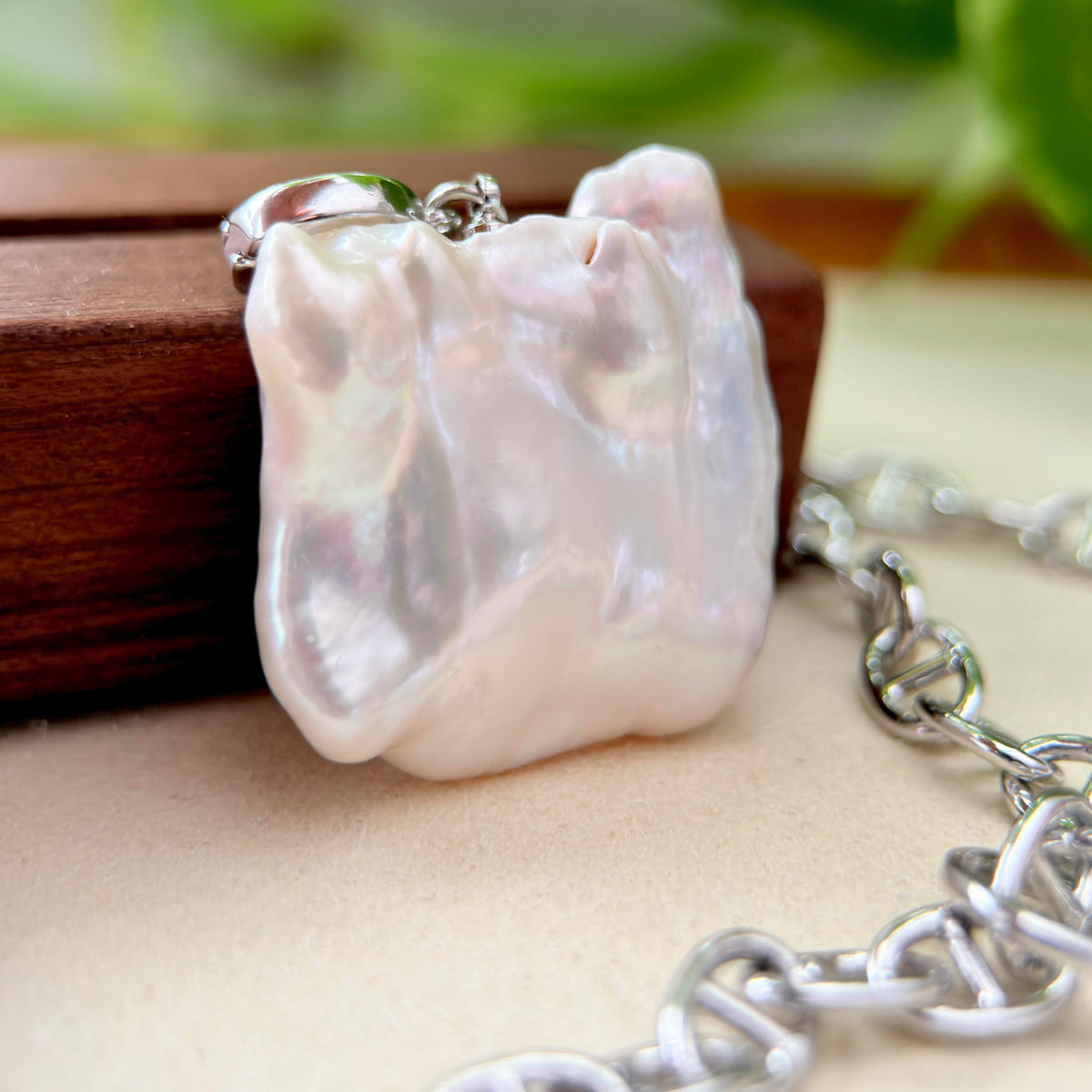 A close-up of a chunky, irregularly shaped white pearl pendant with a silver bail, resting on a dark wooden block with a silver chain draped in the foreground.