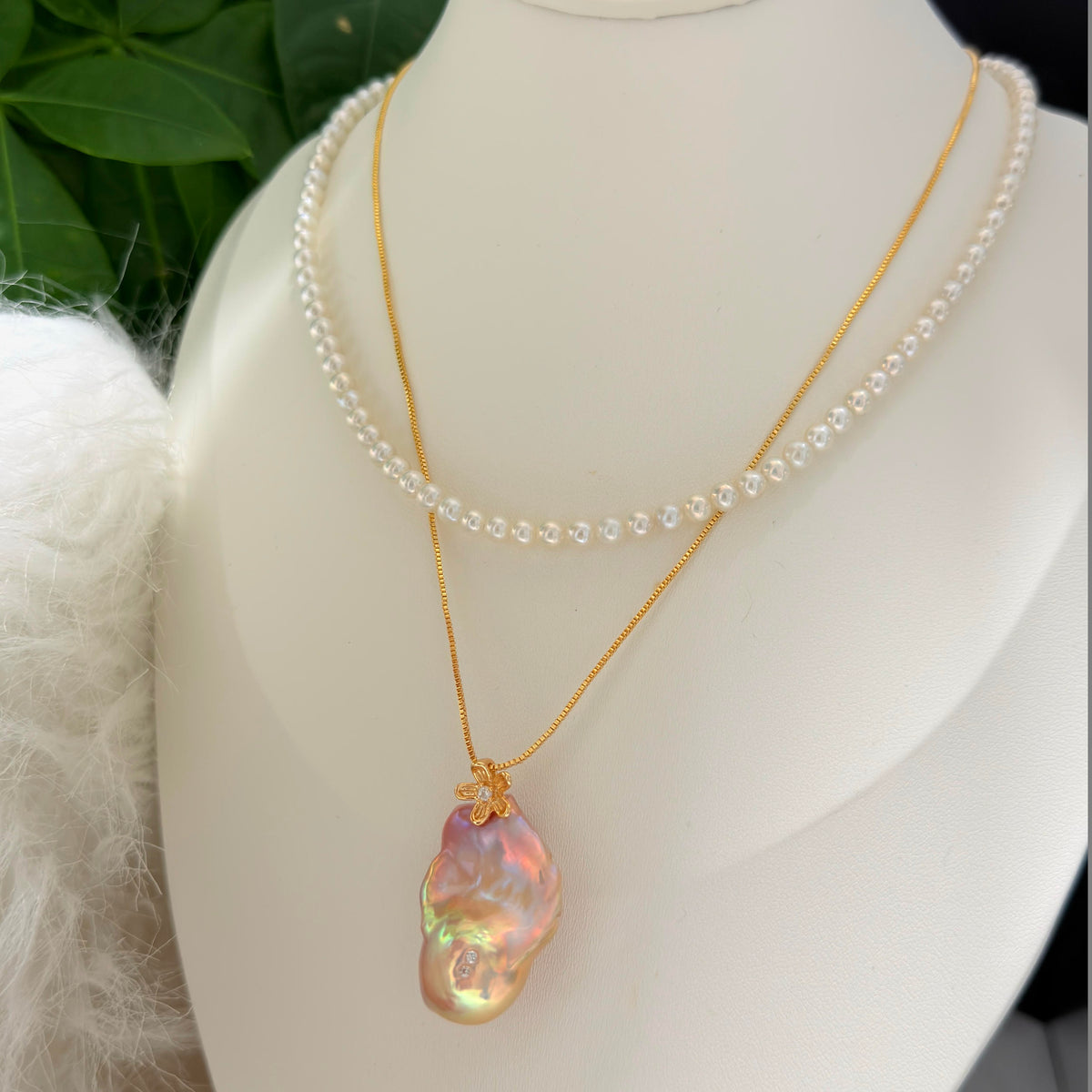 A layered necklace set displayed on a white mannequin bust, featuring a delicate white pearl strand and a gold chain with a large, baroque-shaped iridescent pearl pendant.