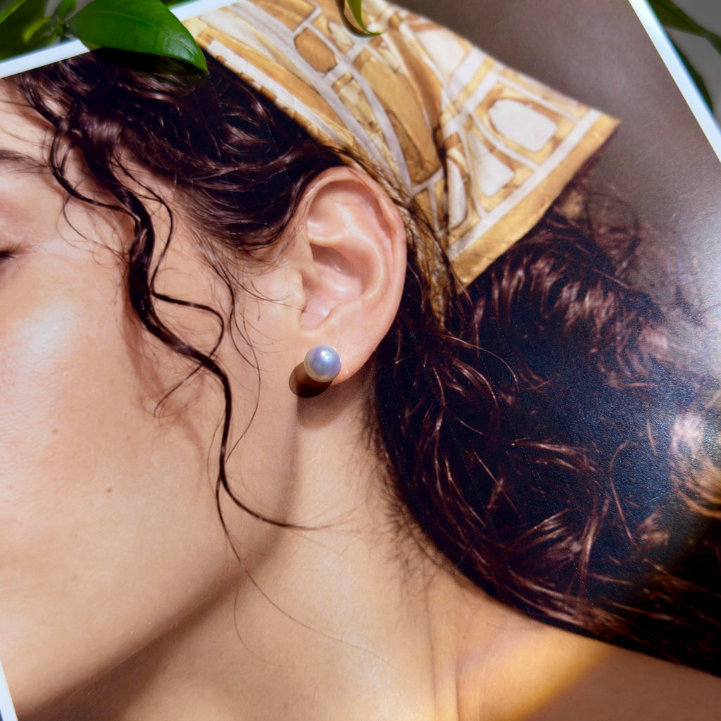 A close-up lifestyle shot of a model wearing a single round pearl stud earring. The model has dark wavy hair and is wearing a gold and white patterned silk headscarf.
