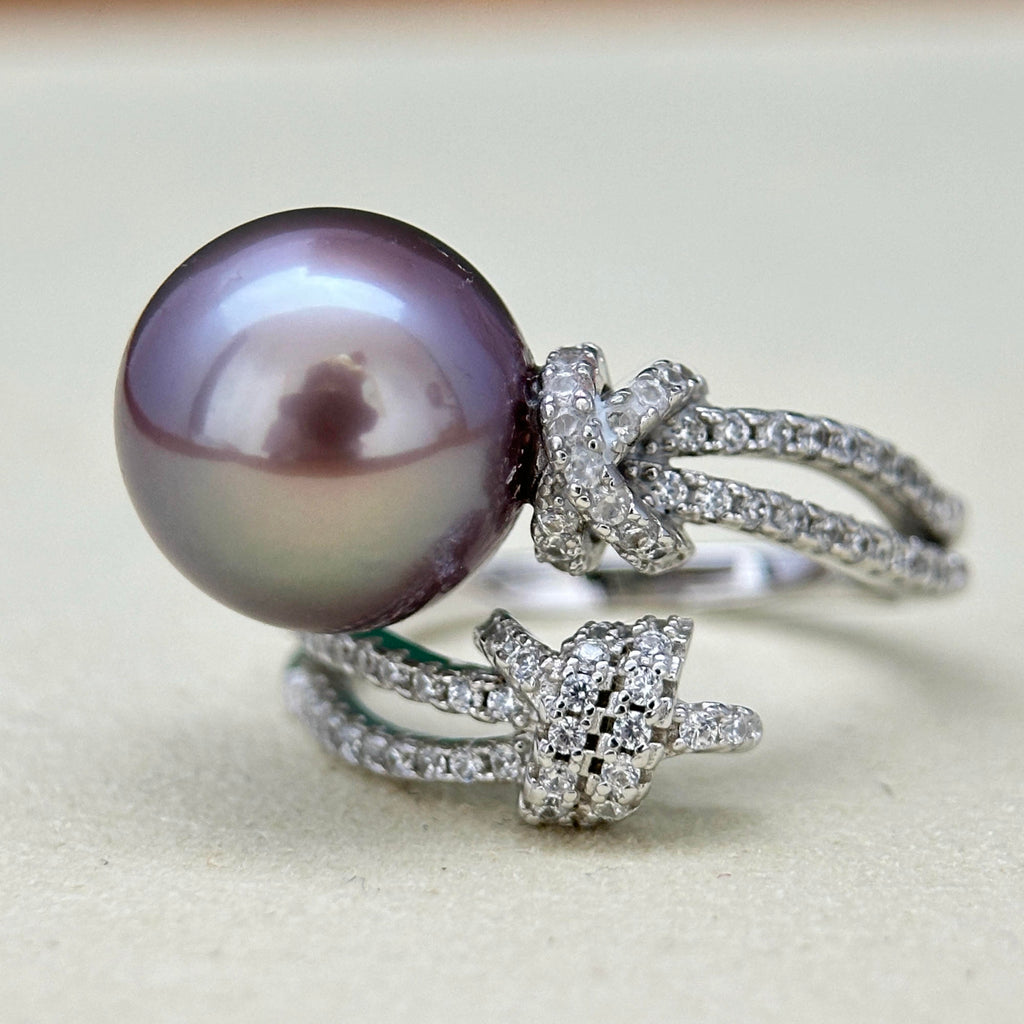 A silver-toned ring featuring a large oval iridescent pearl with a purple-lavender cast, accented by two bow-shaped clusters of small clear stones on a soft beige background.