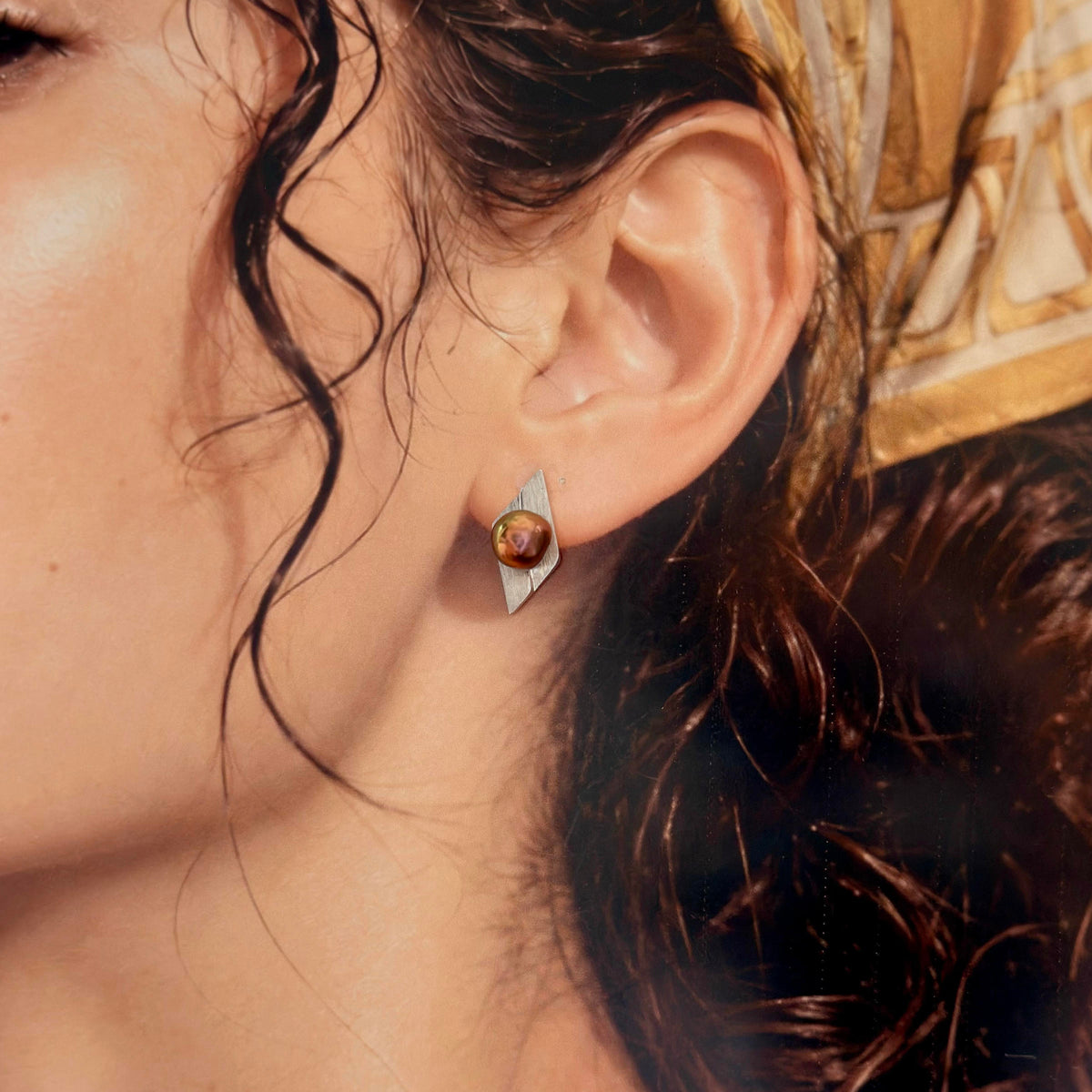 A close-up of a model's ear wearing a silver geometric stud earring with a round amber-brown cabochon stone.