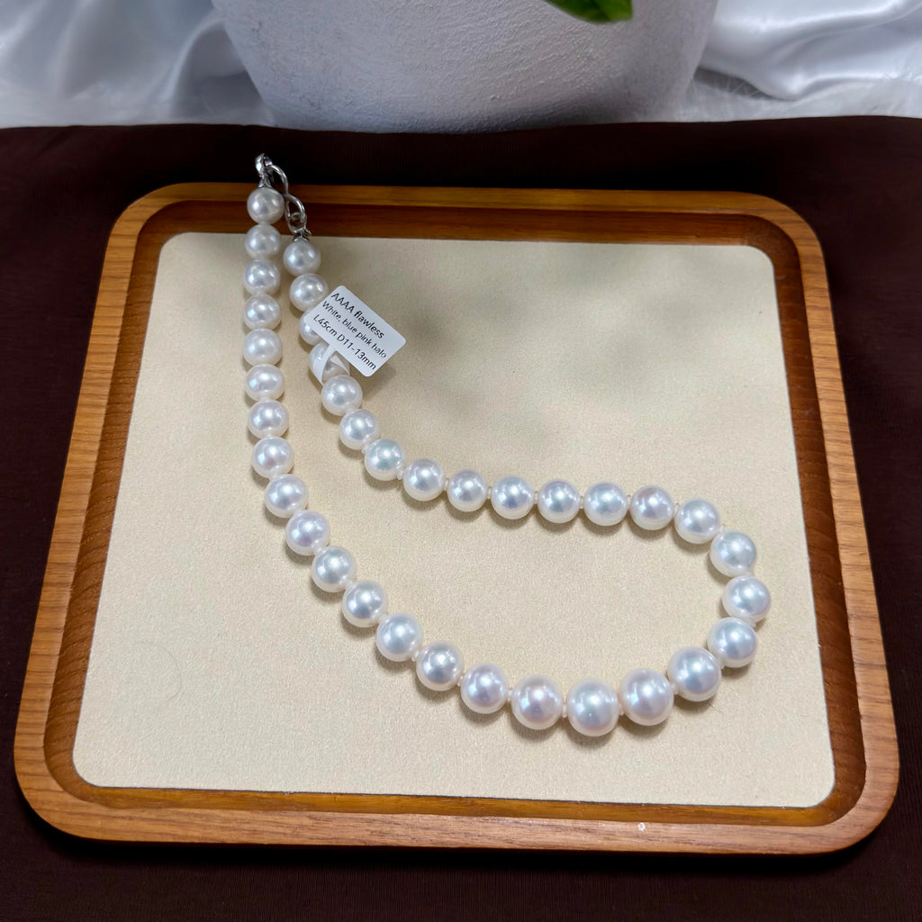 A strand of white freshwater pearls laid out in a U-shape on a beige display mat inside a wooden tray, with a small paper tag attached near the clasp.