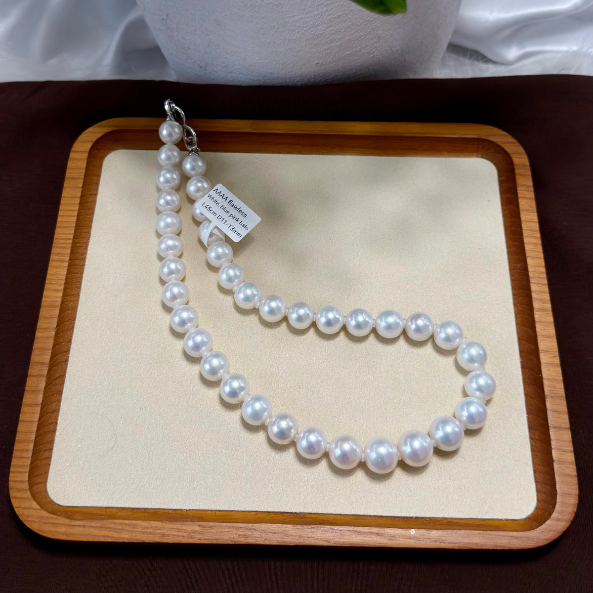 A strand of white freshwater pearls laid out in a U-shape on a beige display mat inside a wooden tray, with a small paper tag attached near the clasp.