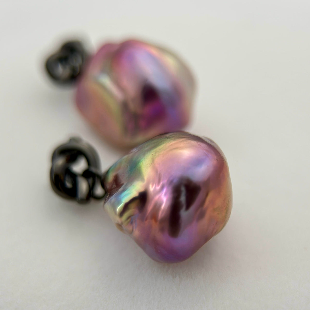 A pair of baroque pearl stud earrings with iridescent purple, pink, and green hues, set in dark metal on a plain white background.