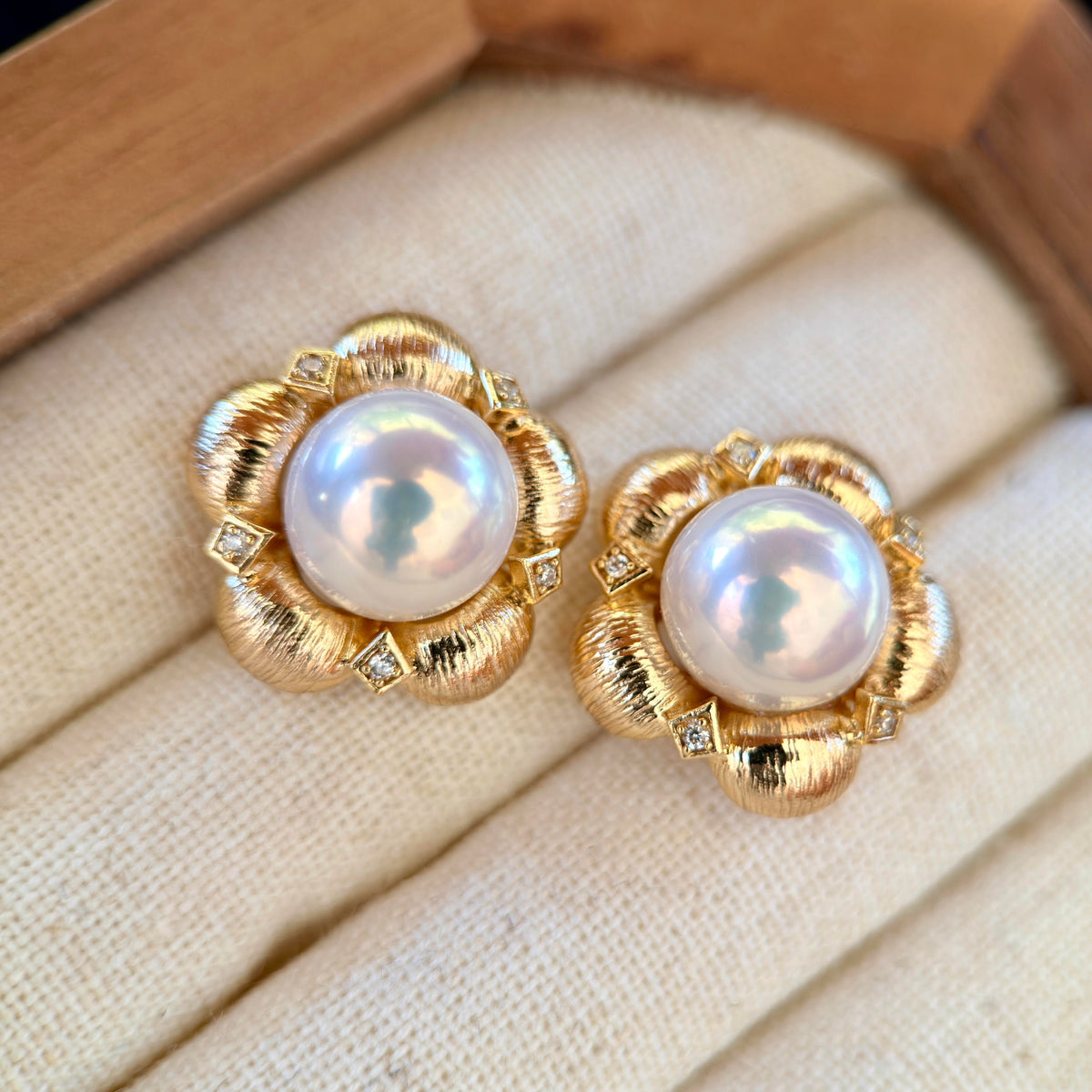 A pair of gold flower-shaped stud earrings with iridescent white pearls and small clear stones, displayed on a cream fabric jewelry cushion.
