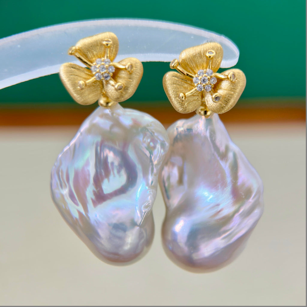 A pair of gold flower-shaped stud earrings with small clear stone centers and large, irregular iridescent pearl-like drops, shown close up against a green and beige background.