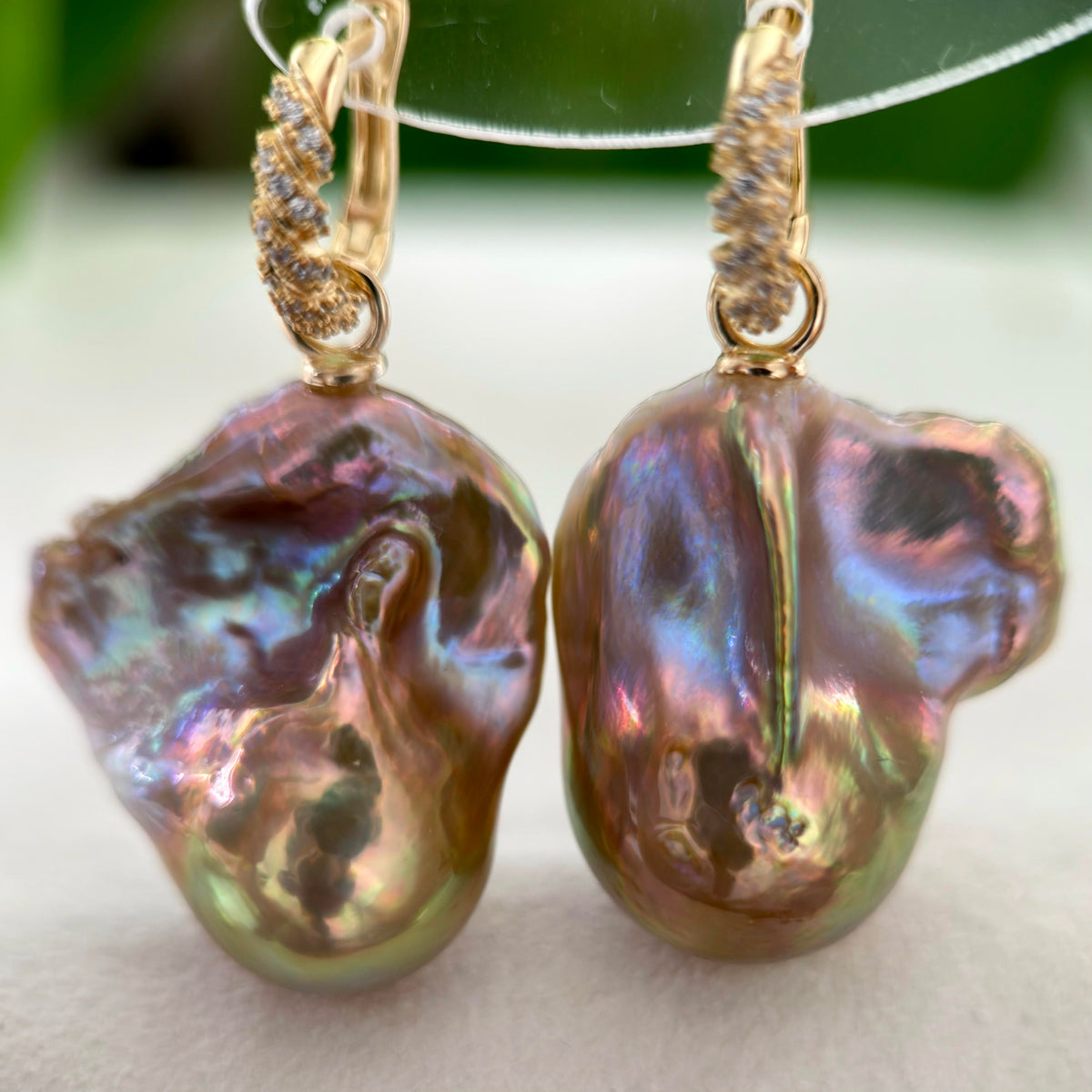 A pair of gold hoop earrings with large, irregular baroque pearls that shimmer with iridescent purple, green, and gold hues, displayed on a clear stand.