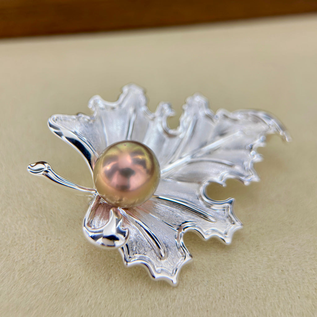 A silver leaf-shaped brooch with a central iridescent pinkish pearl, photographed on a beige surface.