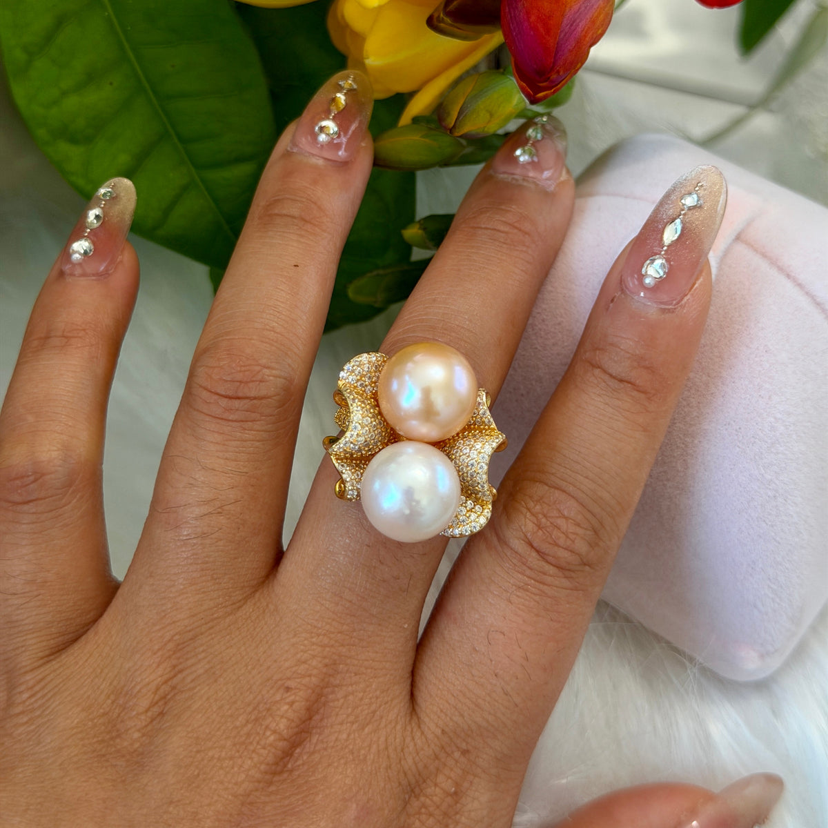 A close-up of a hand wearing a gold ring with two large white pearls, set against a soft white surface with green leaves and yellow flowers in the background.