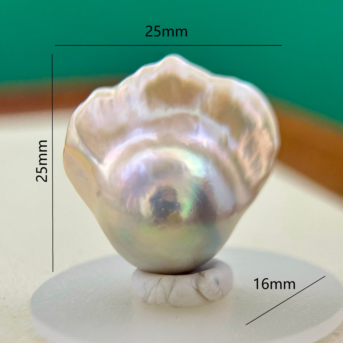 A single iridescent pearl displayed on a small white stand, with measurement lines and labels showing 25mm width, 25mm height, and 16mm base diameter against a blurred green and beige background.