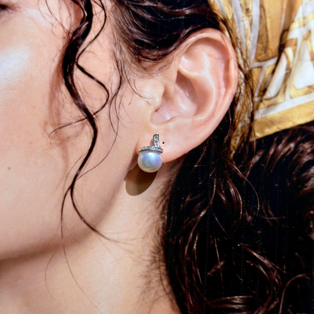 A close-up of a person's ear wearing a single pearl stud earring with a silver-toned setting and small clear accent stones.