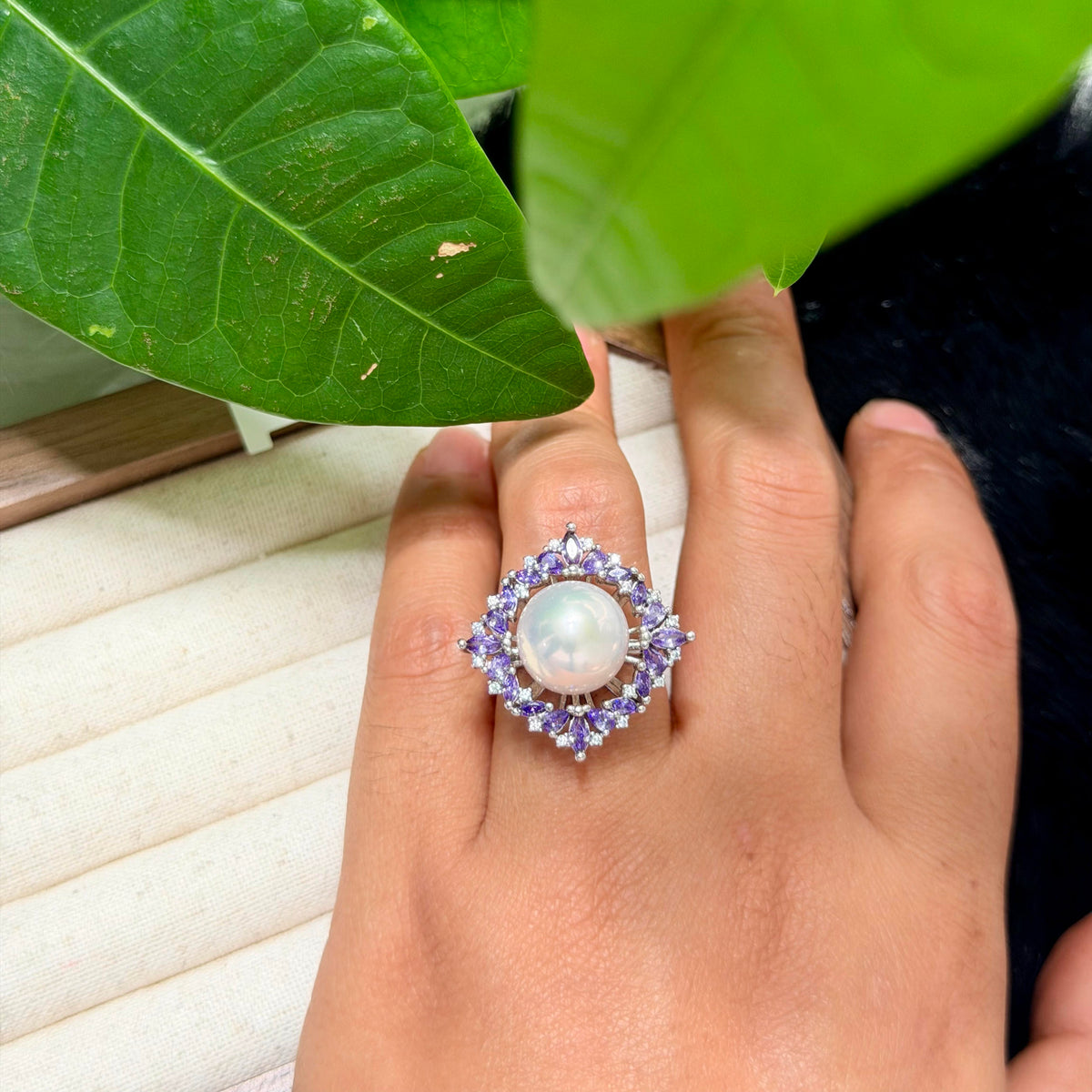 A close-up of a hand wearing a pearl ring with a large white pearl surrounded by a halo of small purple and clear stones, with green leaves in the foreground.